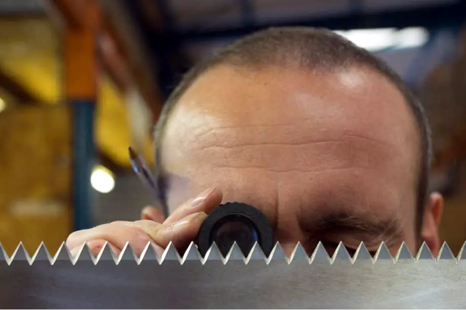 A large steel, saw-toothed, industrial machine knife being inspected closely by an engineer with a magnifying glass